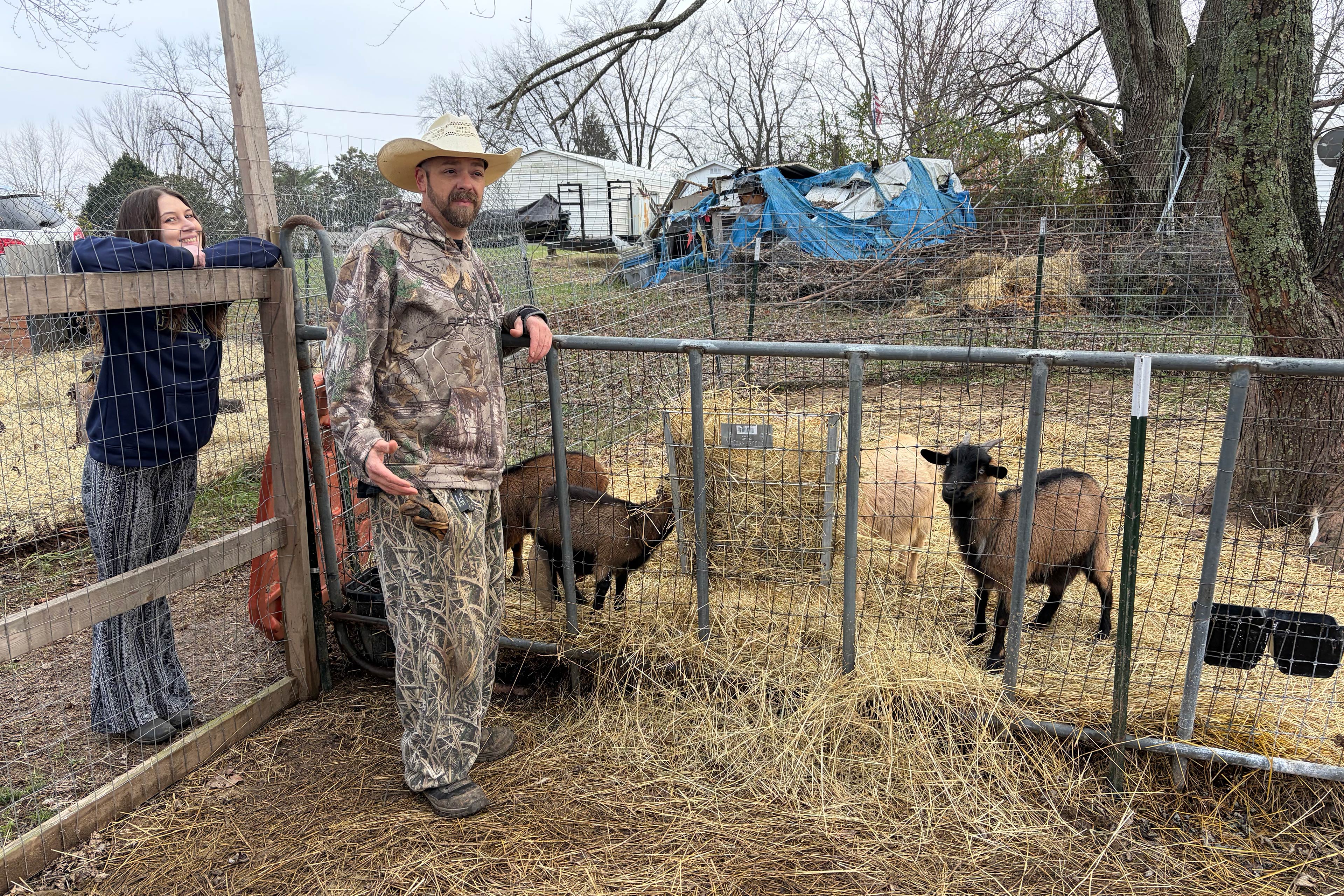 A man and a woman lean against the fences of a fenced-in area with straw on the ground and four visible goats. The woman with straight dark hair wears a dark blue sweatshirt with striped pants and smiles at the camera. The man with a beard wears a straw hat, camouflage sweatshirt, and camouflage pants is in the middle of talking and looks a something off-camera.